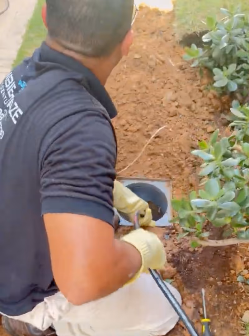 Homem de luvas amarelas trabalhando na manutenção de um encanamento enterrado próximo a uma planta.